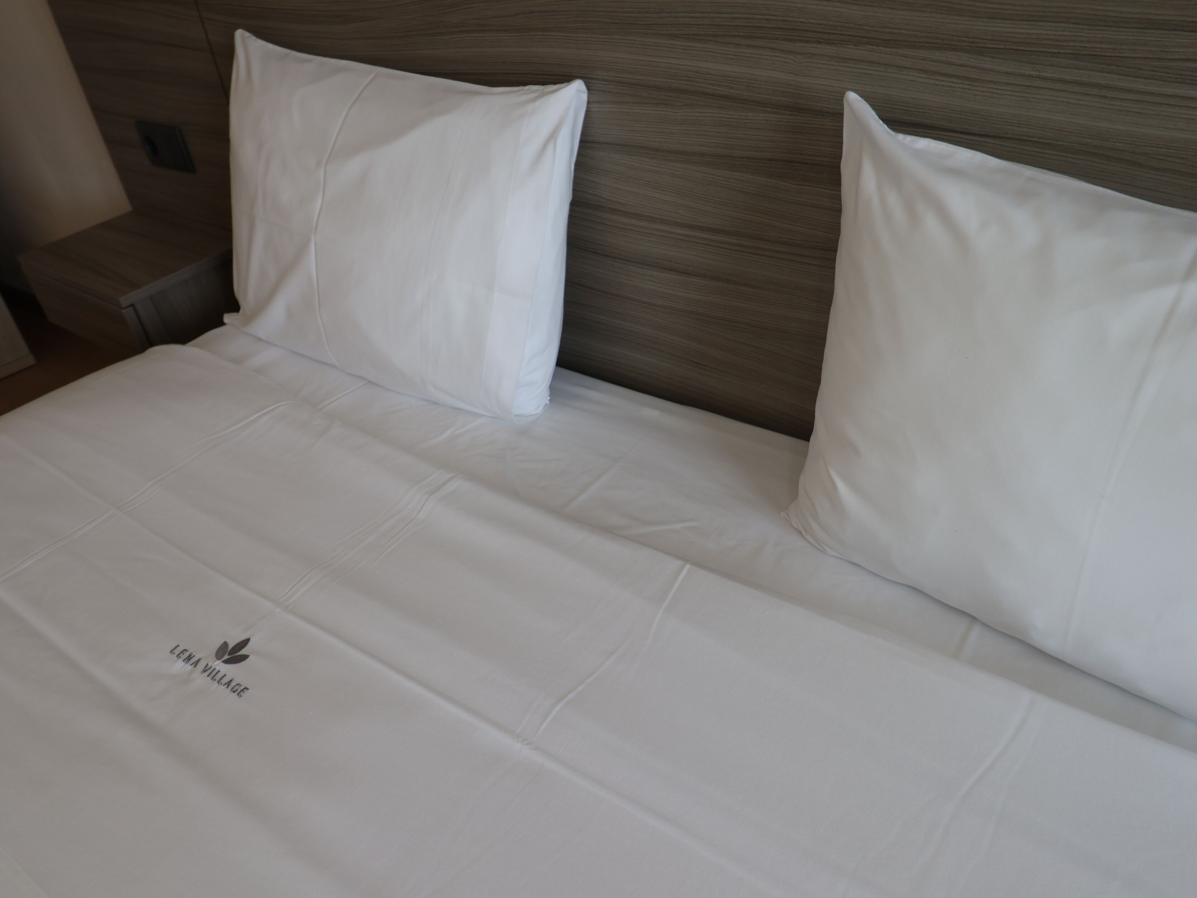 Una cama bien tendida con sábanas y almohadas blancas, un cabecero de madera y un logotipo en la colcha.