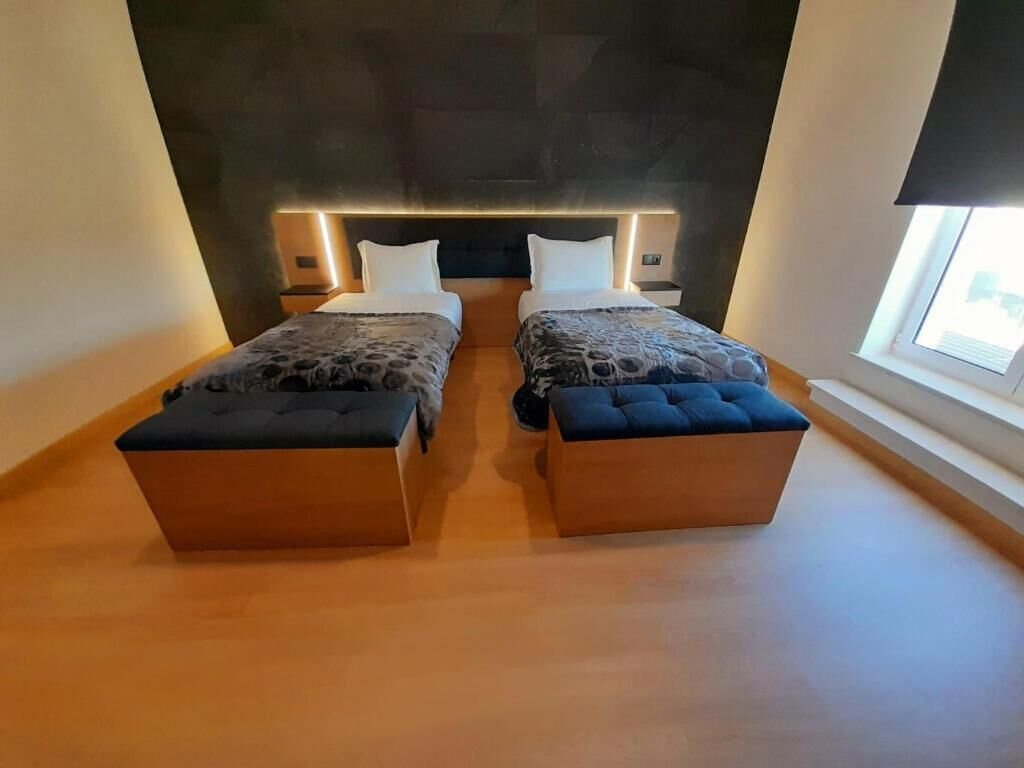 A modern bedroom with two beds, a dark headboard, bedside tables, and a window with a view.