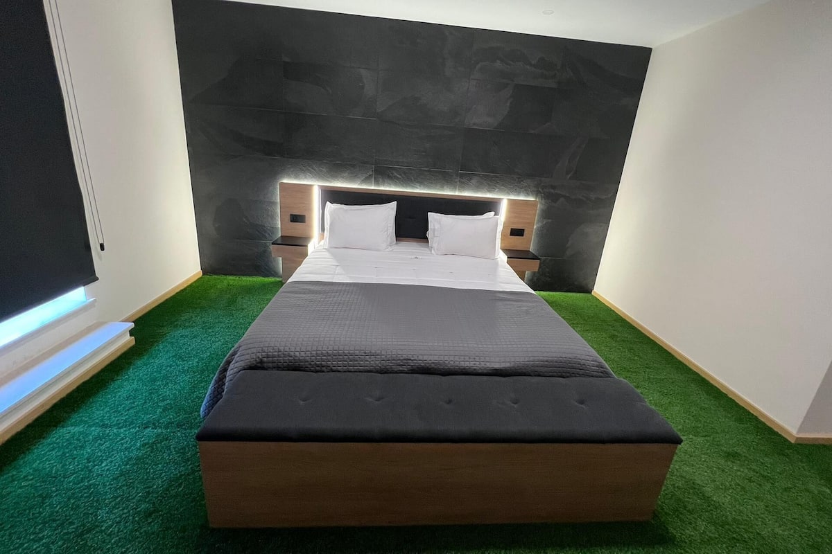 A modern hotel room with a large bed, green carpet, and a dark wall.