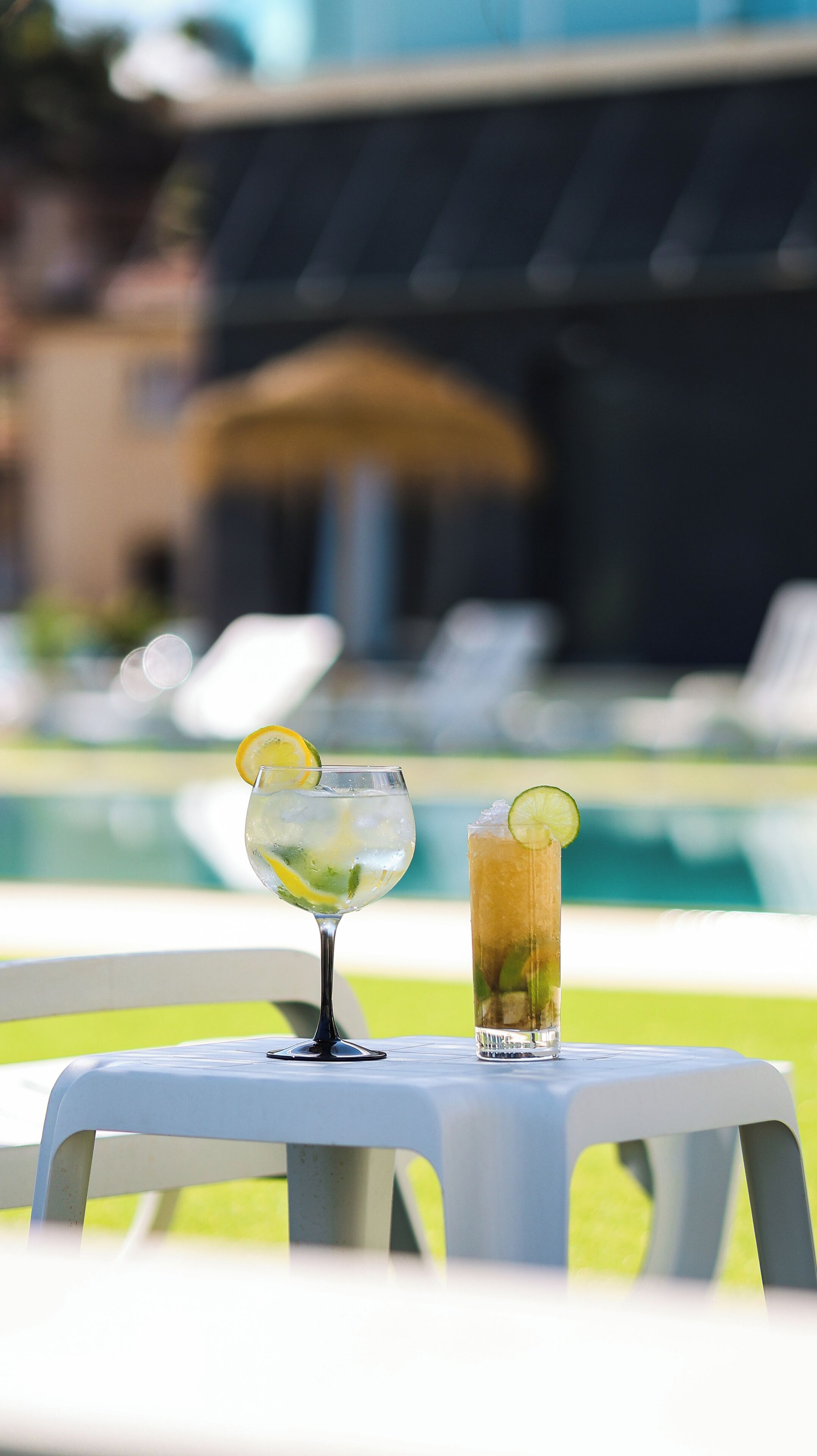 Two glasses of beverages on a table by a poolside, with a blurred background of a pool and lounge chairs.