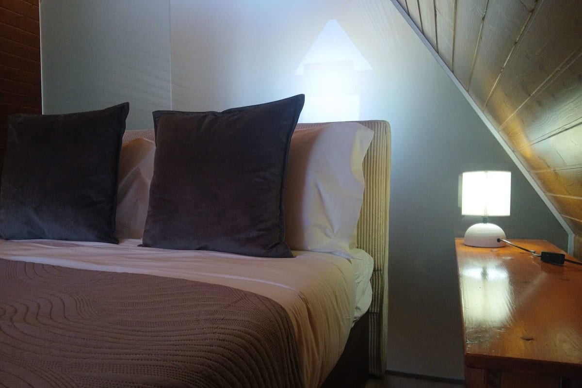 A neatly made bed with dark pillows and a light-colored bedspread, a bedside table with a lamp, and a wooden headboard.