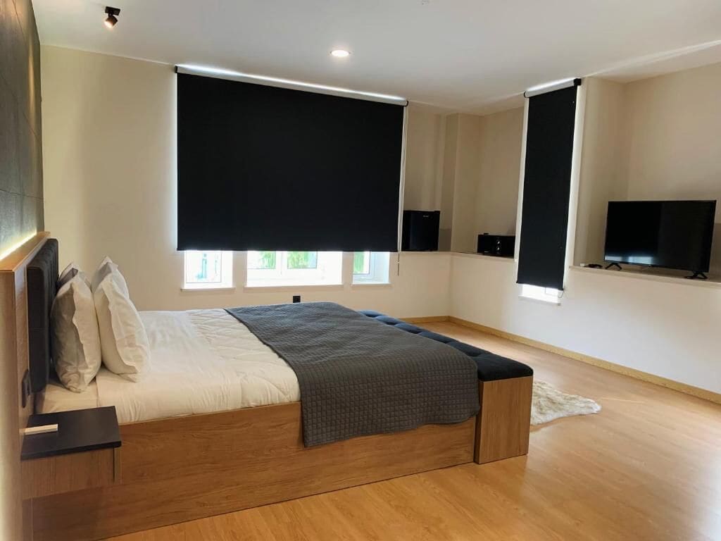 A modern bedroom with a wooden bed, blackout curtains, a flat-screen TV, and a small rug.