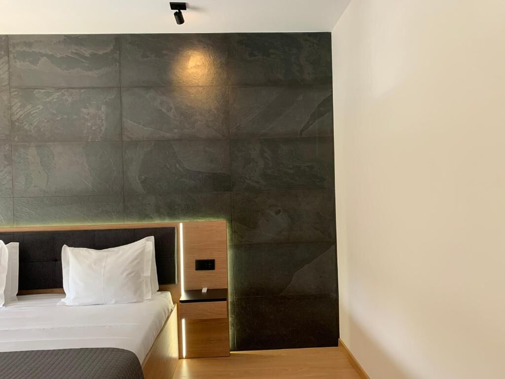 A modern hotel room with a large bed, a wooden headboard, a nightstand, and a dark textured wall.