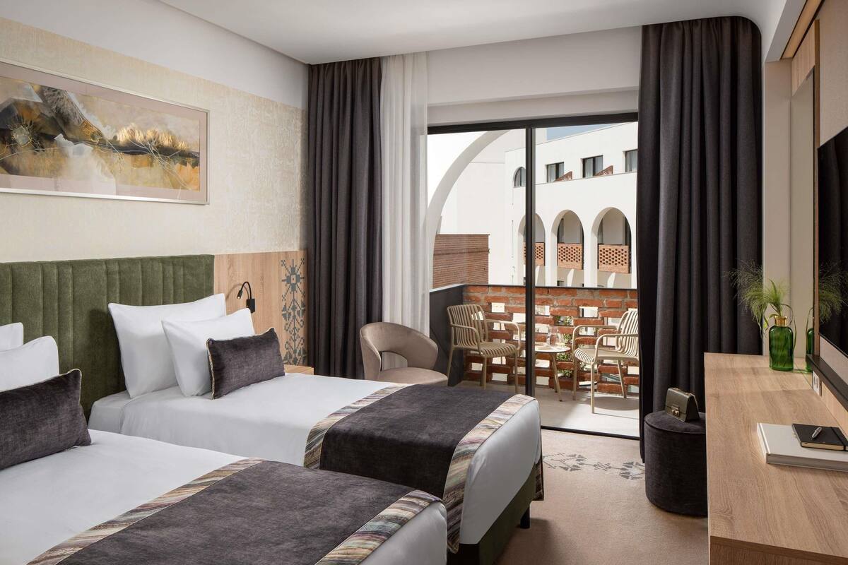 Une chambre d’hôtel avec deux lits, un bureau, une chaise et un balcon donnant sur des immeubles.