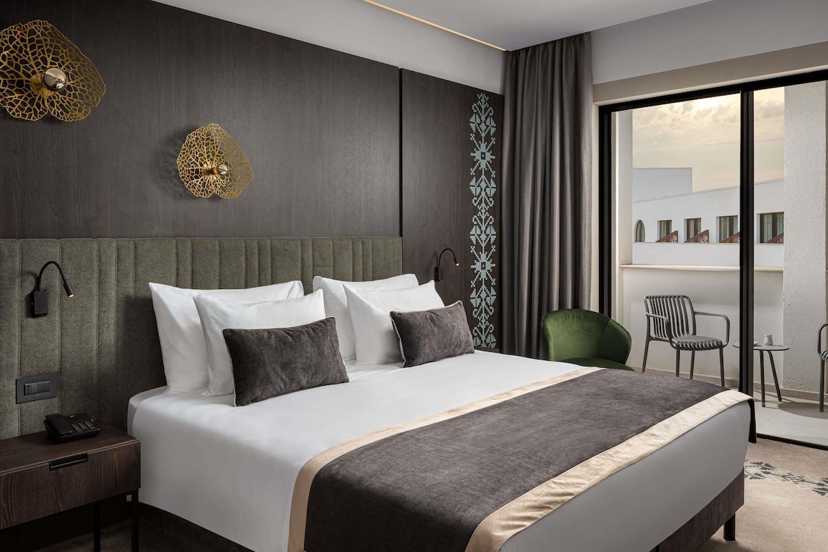 Une chambre d’hôtel moderne dotée d’un grand lit, d’une table de chevet, d’un bureau et offrant une vue sur la ville grâce à de grandes fenêtres.