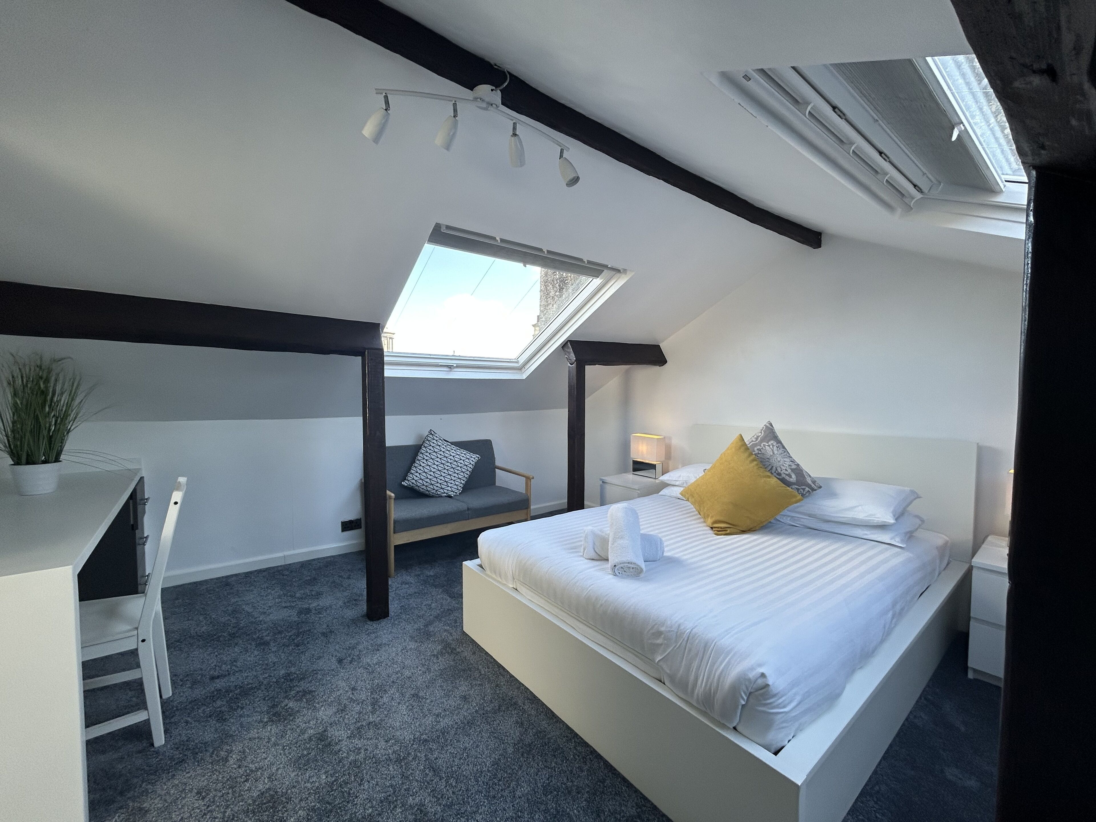 A modern bedroom with a skylight, a bed with white linens, a bedside table, a desk, and a chair.