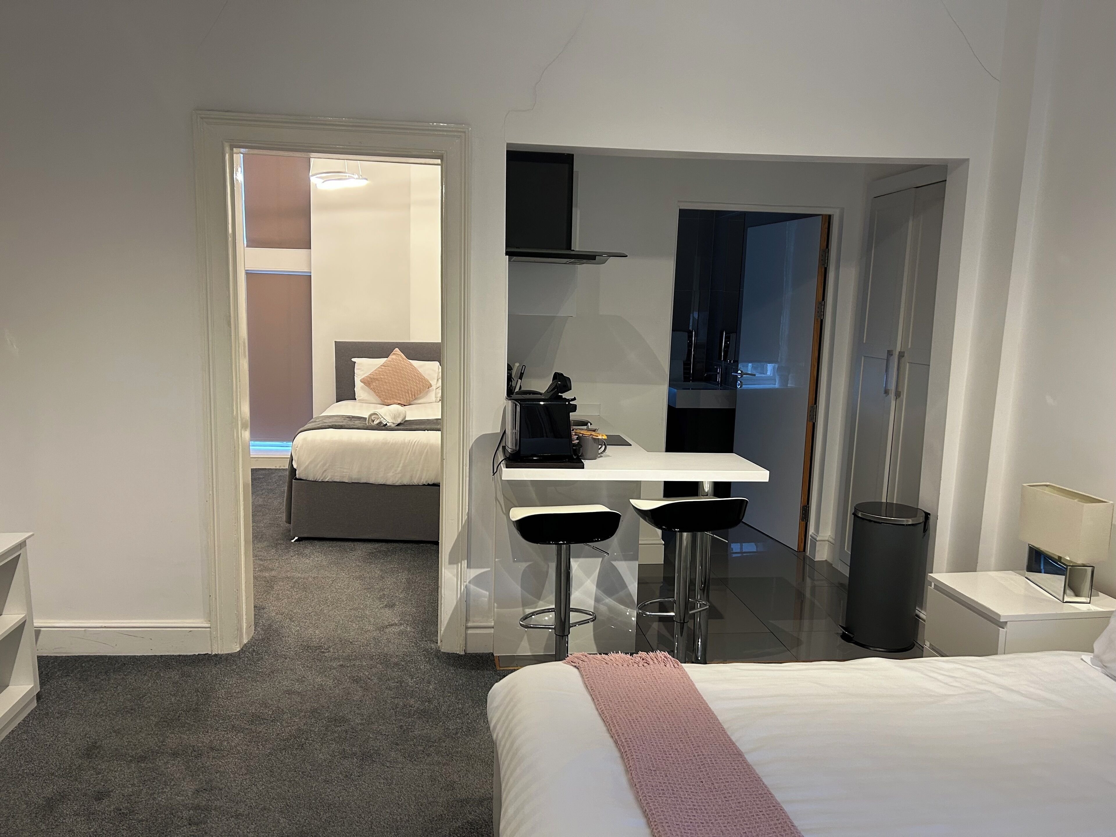 A modern hotel room with a bed, a desk with two chairs, and a small kitchenette.