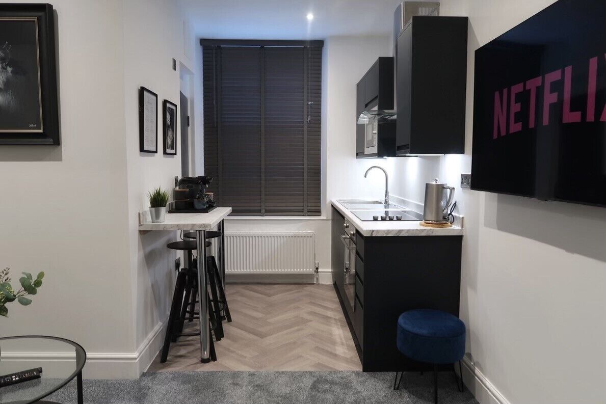 A compact kitchen with a black countertop, a sink, and a coffee machine. There is a blue stool and a framed picture on the wall.
