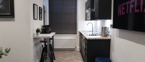 A compact kitchen with a black countertop, a sink, and a coffee machine. There is a blue stool and a framed picture on the wall.
