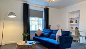 A modern living room with a blue sofa, a round coffee table, a floor lamp, and a bookshelf. - Sun Street ApartHotel (Lancaster)