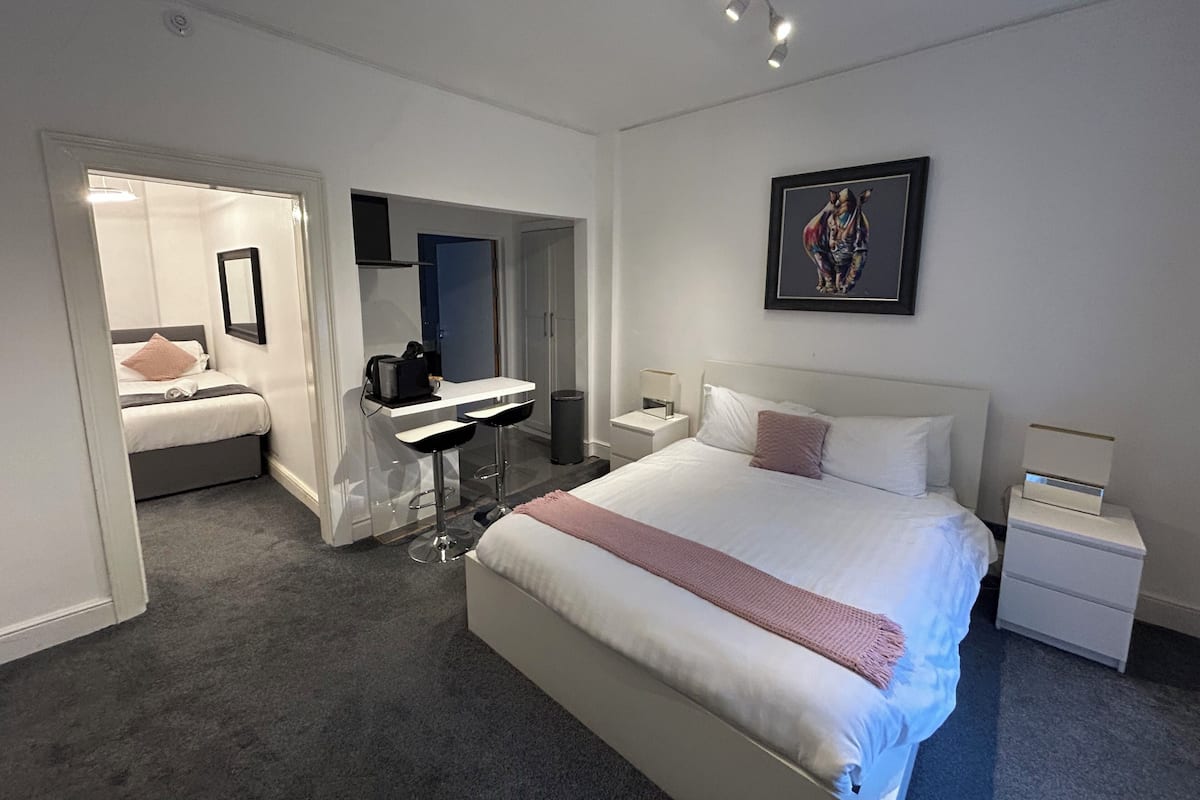 A modern hotel room with a large bed, a desk, and a mirror. There is a framed artwork on the wall and a small nightstand with a lamp.