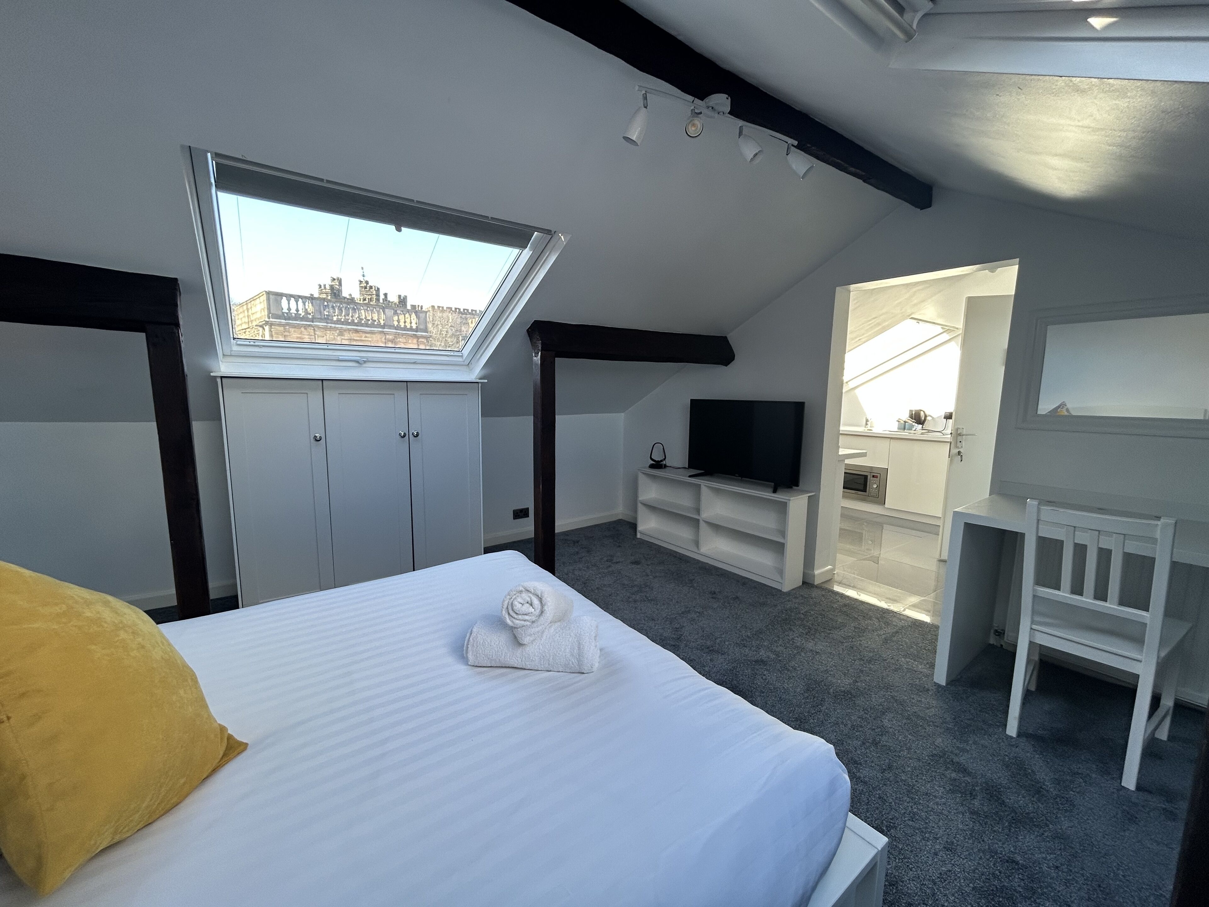 A bedroom with a skylight, a bed with a white comforter and a yellow pillow, a TV on a white stand, and a bathroom visible through an open door.