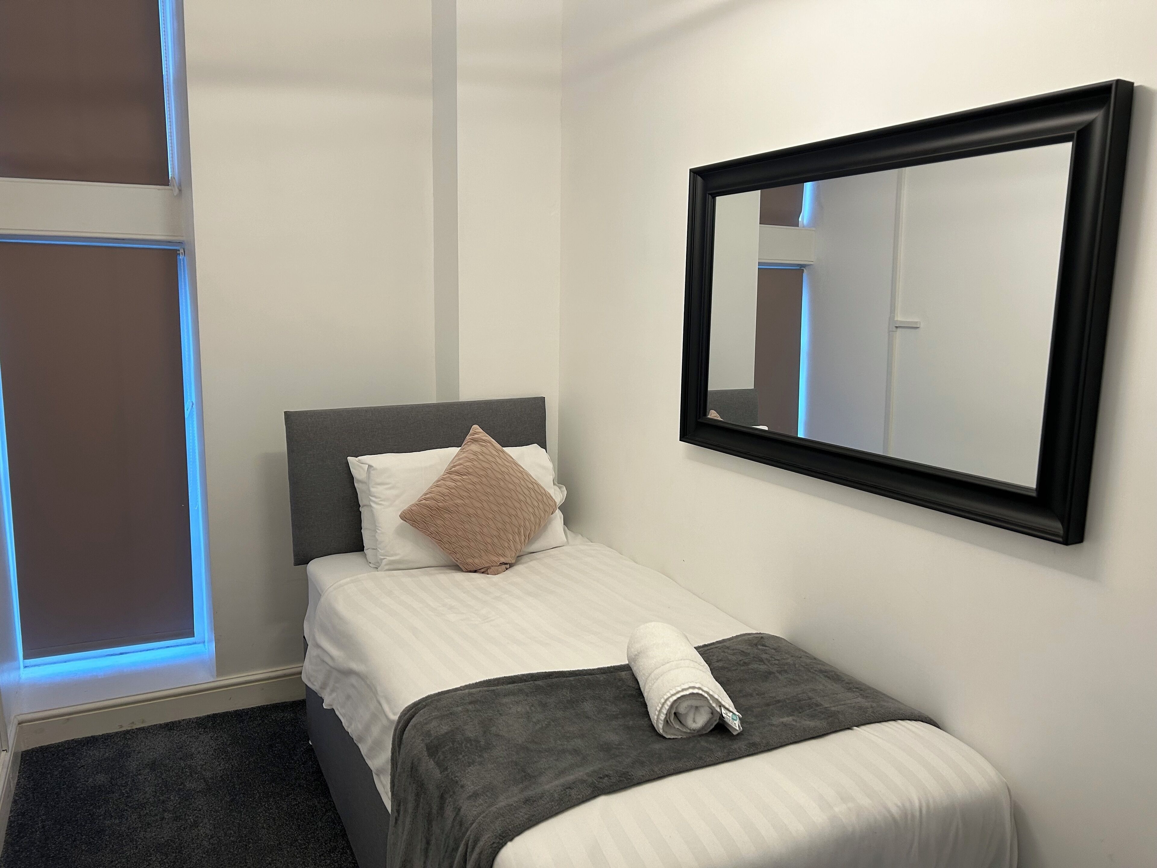 Un lit bien fait, avec une couverture grise et une serviette enroulée, un oreiller beige et un grand miroir accroché au mur.