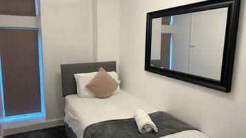 Una cama bien hecha con una manta gris, una toalla enrollada, un cojín color beige y un espejo grande en la pared.