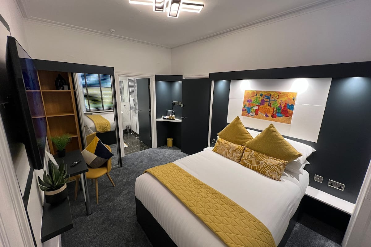 A hotel room with a bed, a desk, a chair, a plant, and a painting on the wall.