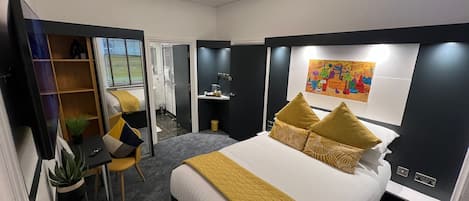 A modern hotel room with a large bed, a desk, a chair, a small plant, and a painting on the wall.