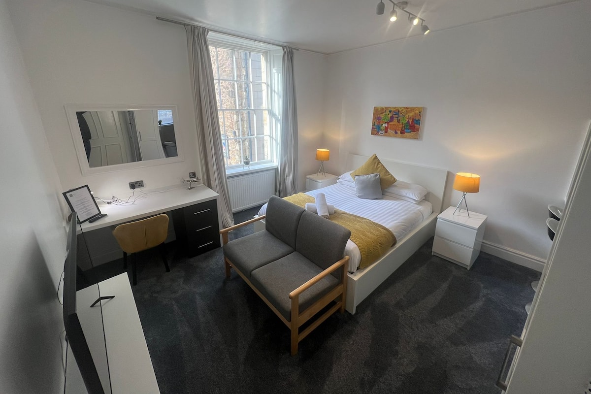 A modern hotel room with a bed, bedside tables, a desk, a chair, and a large window.