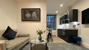 A modern living room with a sofa, a coffee table, and a painting. There is a kitchen area with a microwave and cabinets. - Sun Street ApartHotel (Lancaster)