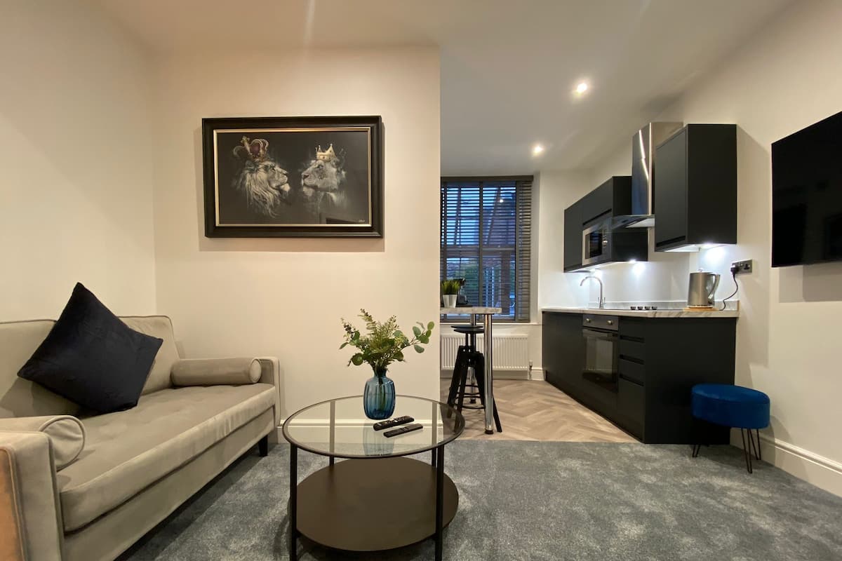 A modern living room with a sofa, a coffee table, and a painting. There is a kitchen area with a microwave and cabinets.