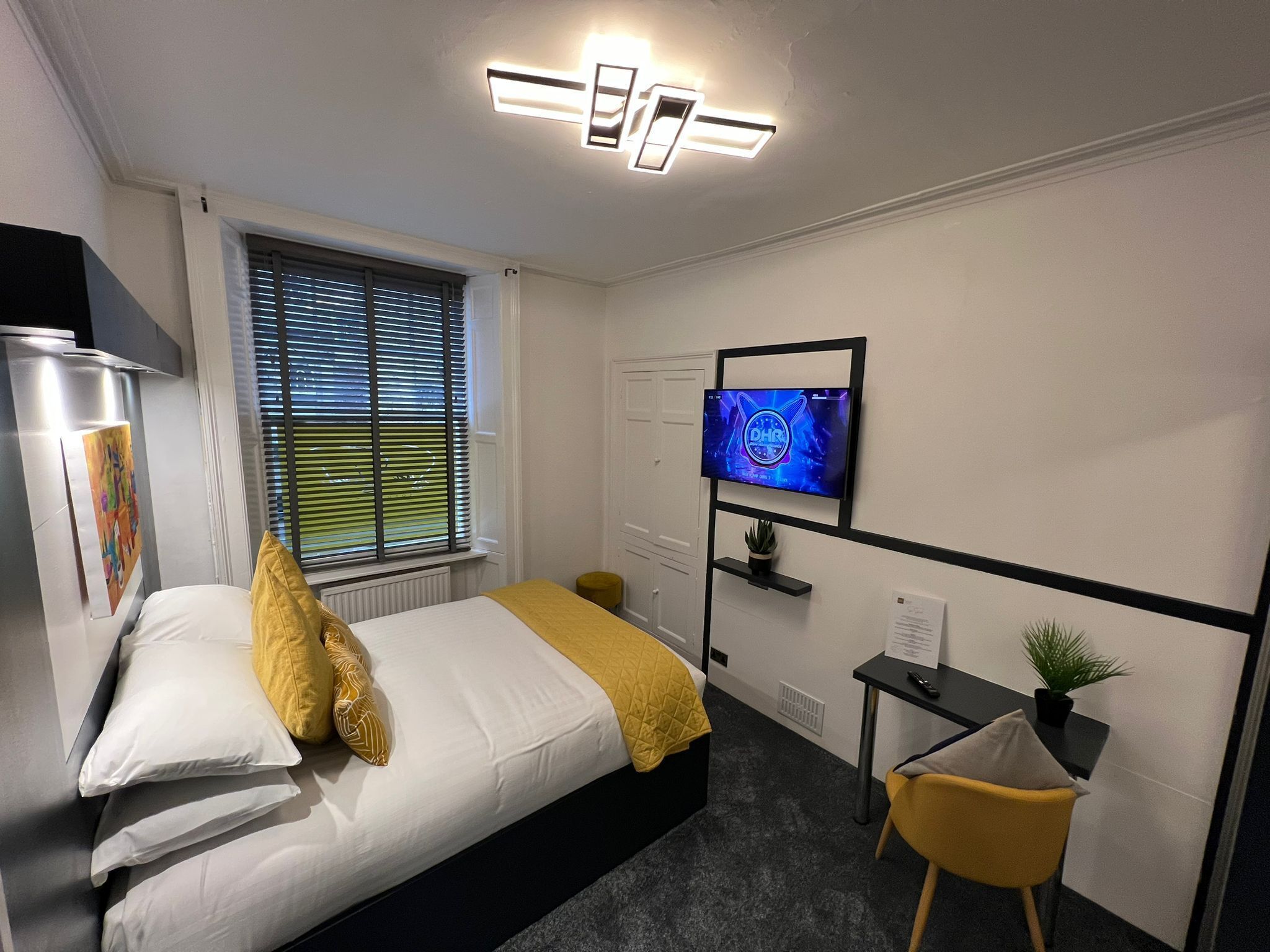 A modern hotel room with a bed, a TV mounted on the wall, a small desk with a plant, and a window with blinds.
