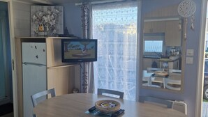 Un comedor con una mesa redonda y cuatro sillas, un refrigerador, un televisor y una cocina visible a través de una puerta corrediza.