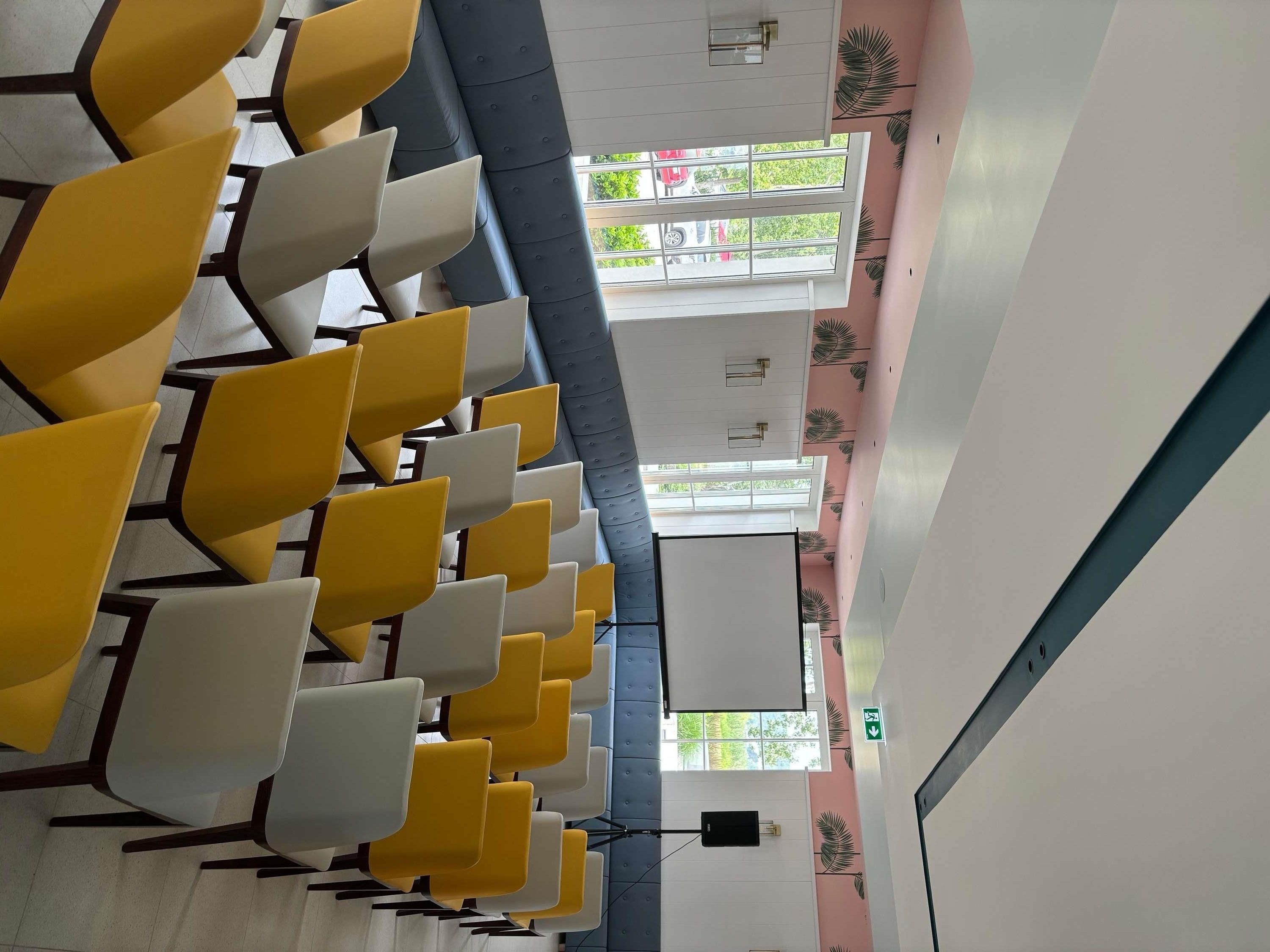 A row of chairs with a high ceiling and windows, a wall with a leaf pattern, and a green exit sign.