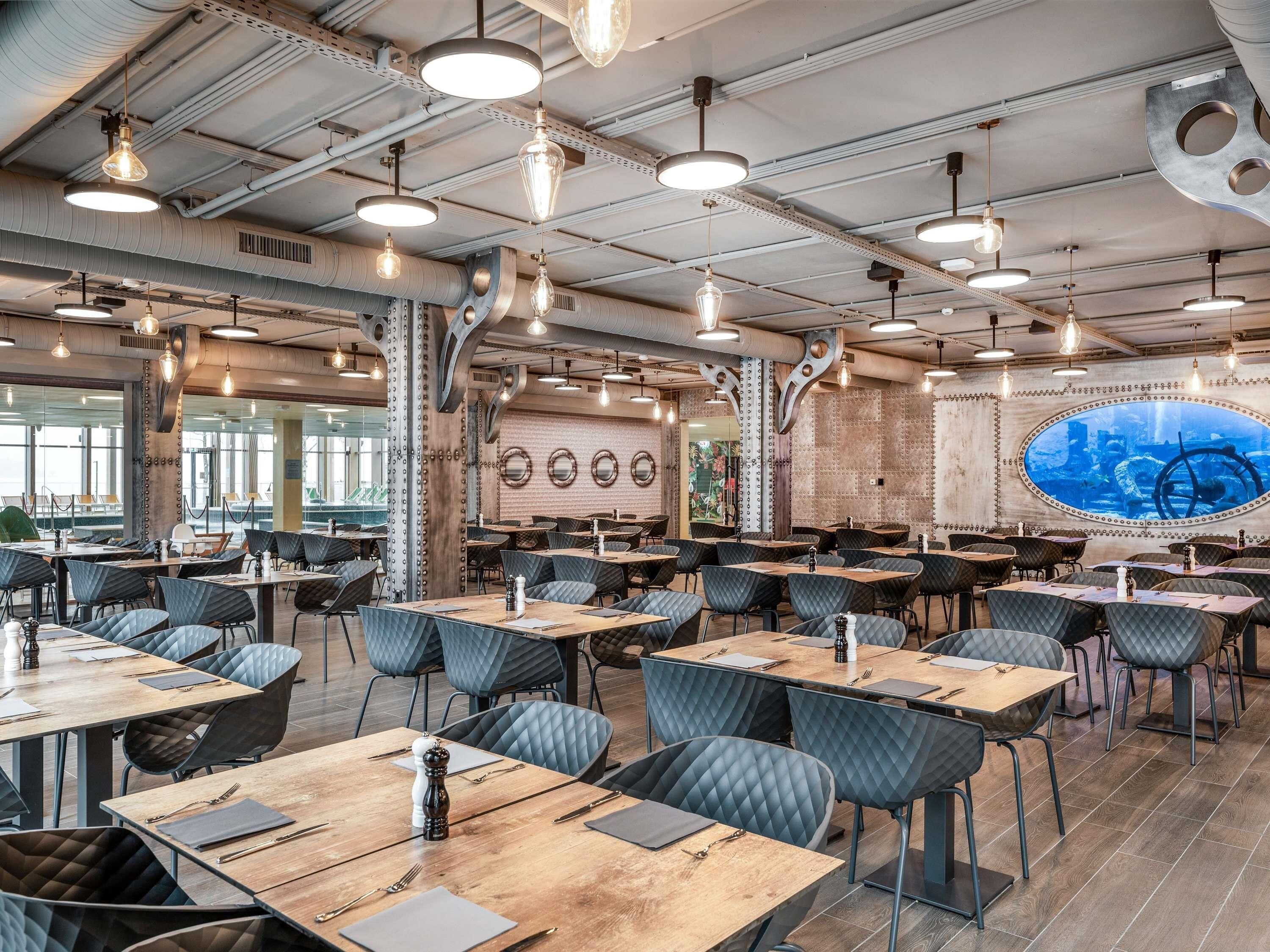 A modern restaurant with high ceilings, exposed ductwork, and industrial-style lighting. Tables are arranged in a spacious layout with cushioned chairs.