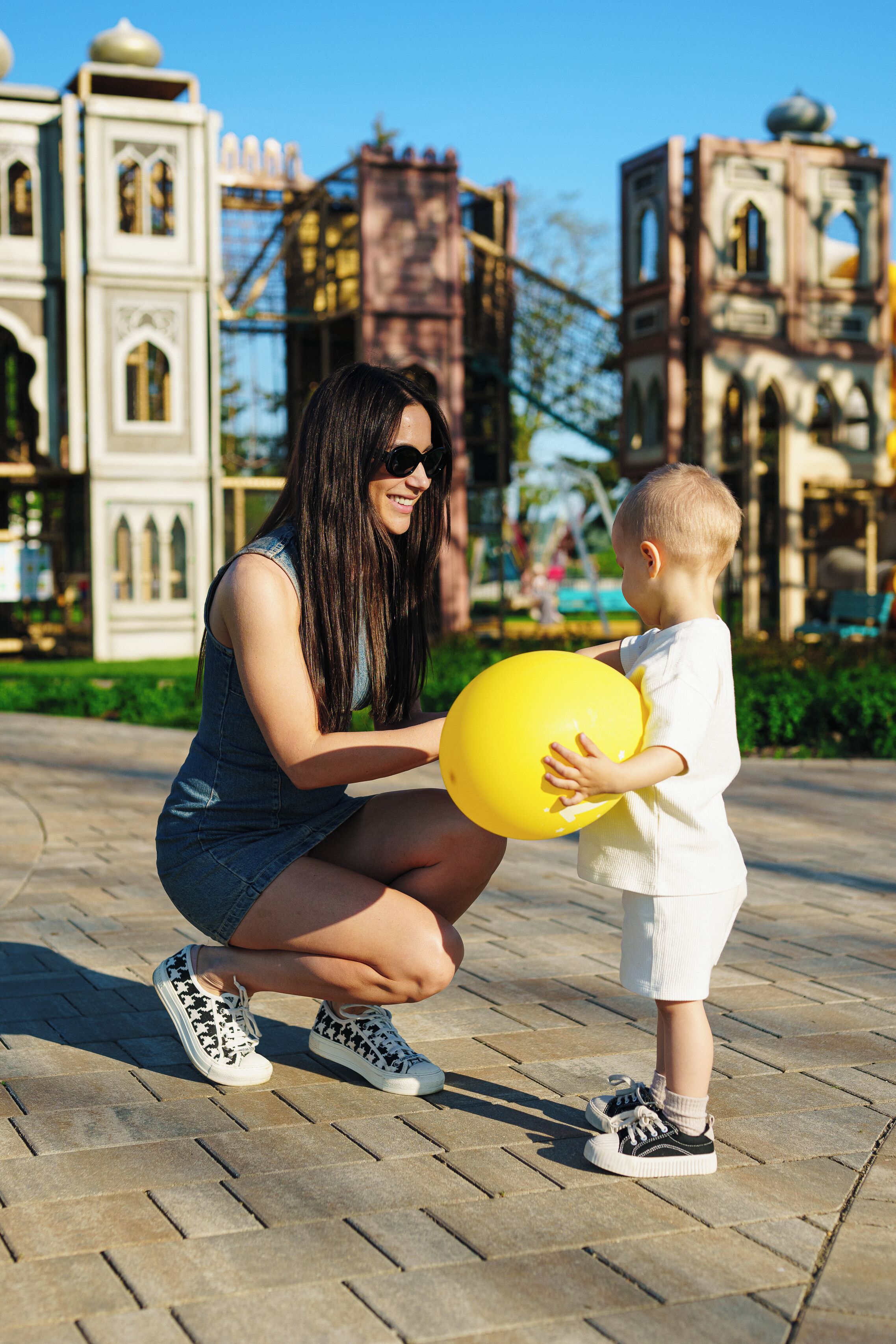 Eine Frau und ein Kind halten einen gelben Ball, im Hintergrund sind architektonische Gebäude zu sehen.