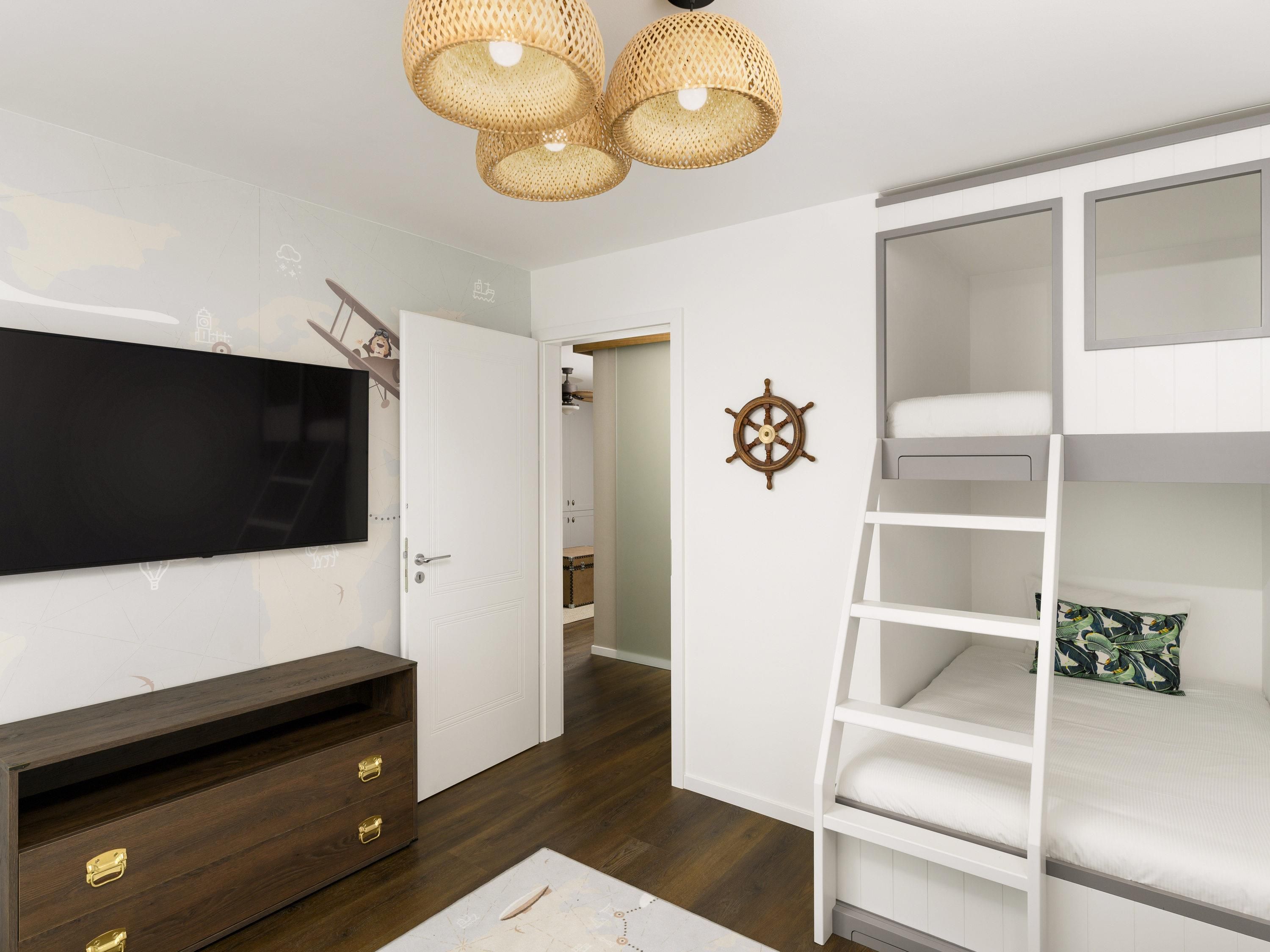 Quarto com cama de solteiro, televisão, cómoda de madeira e roda de navio fixada na parede.