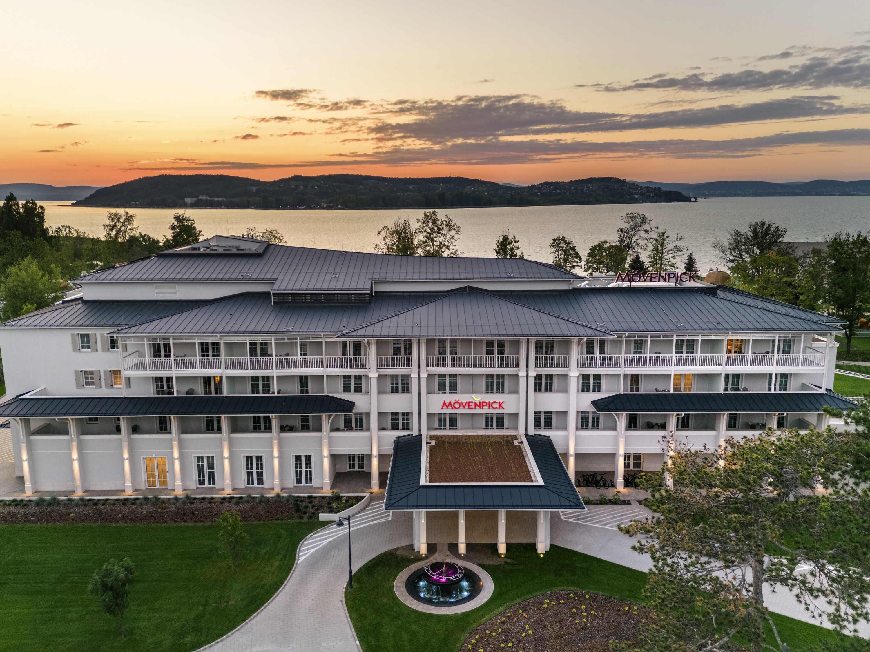 A large, multi-story hotel with a prominent 'Mövenpick' sign, a central courtyard, and a view of a lake at sunset.