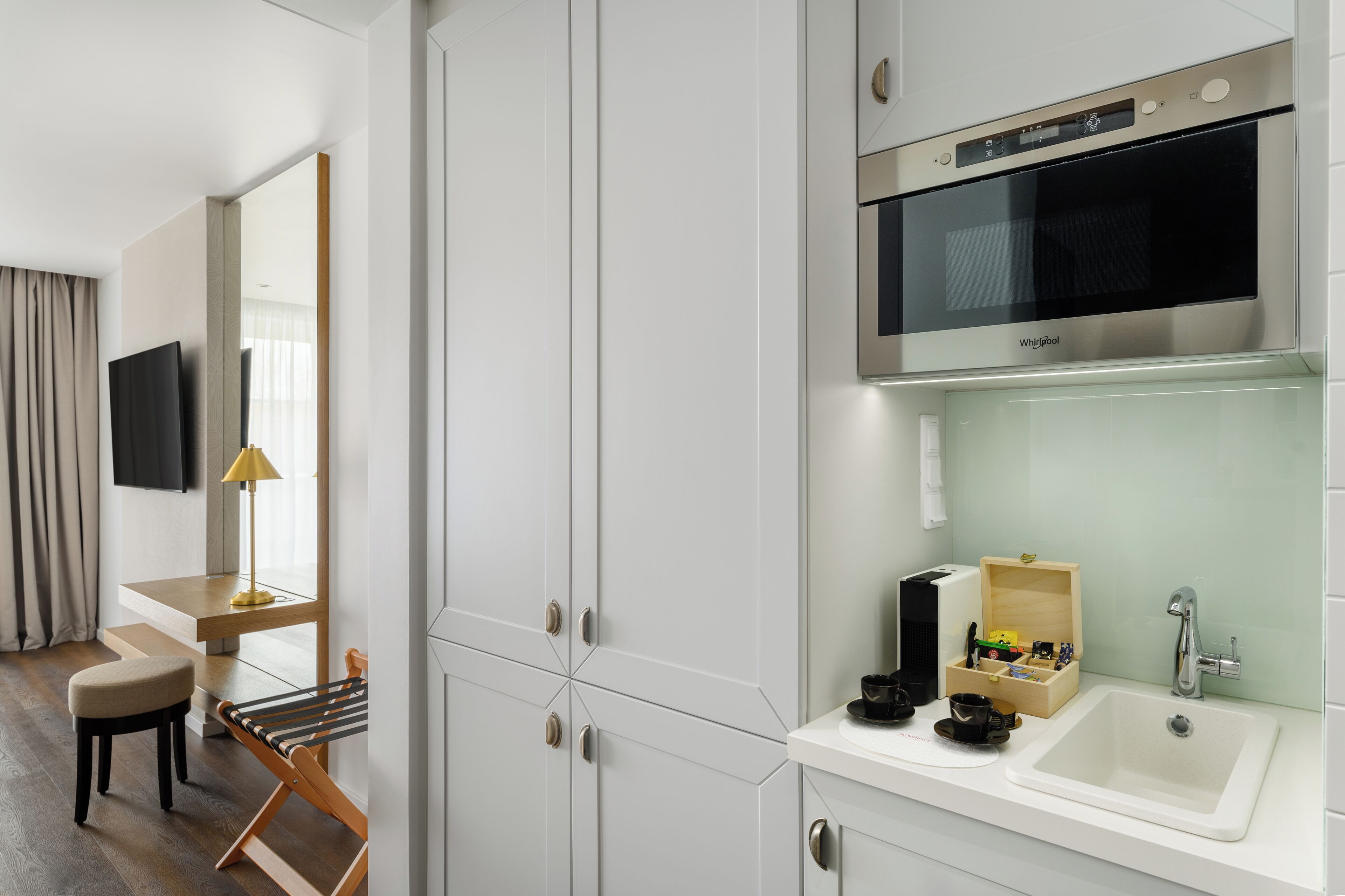Ein modernes Hotelzimmer mit Küchenzeile, einem Flachbildfernseher, einer hölzernen Bank und einem Schreibtisch mit Lampe.