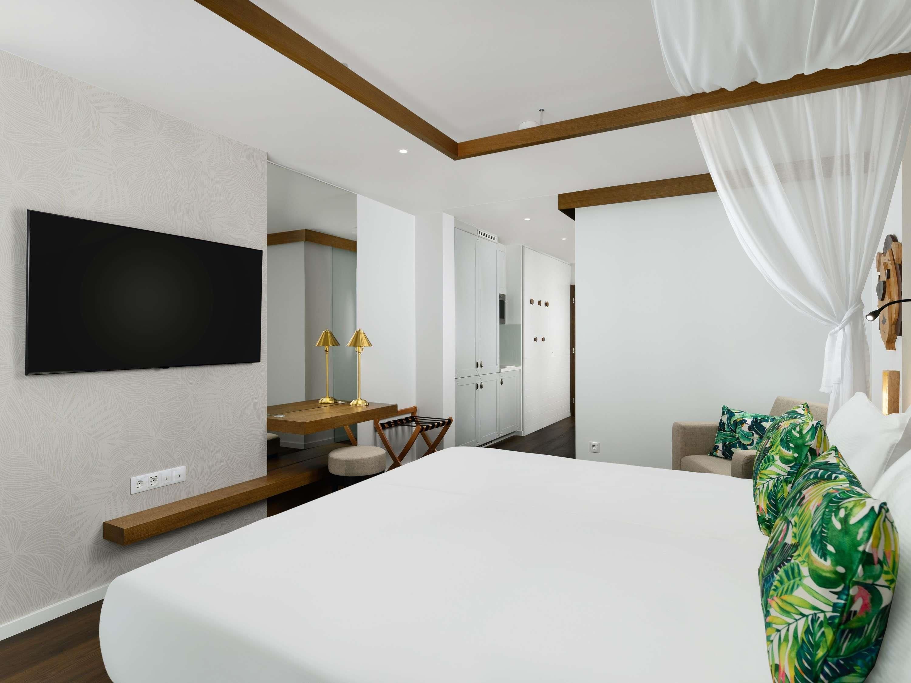 Una habitación de hotel moderna con una cama grande, un televisor de pantalla plana montado en la pared, un escritorio de madera con una silla y un espejo amplio.
