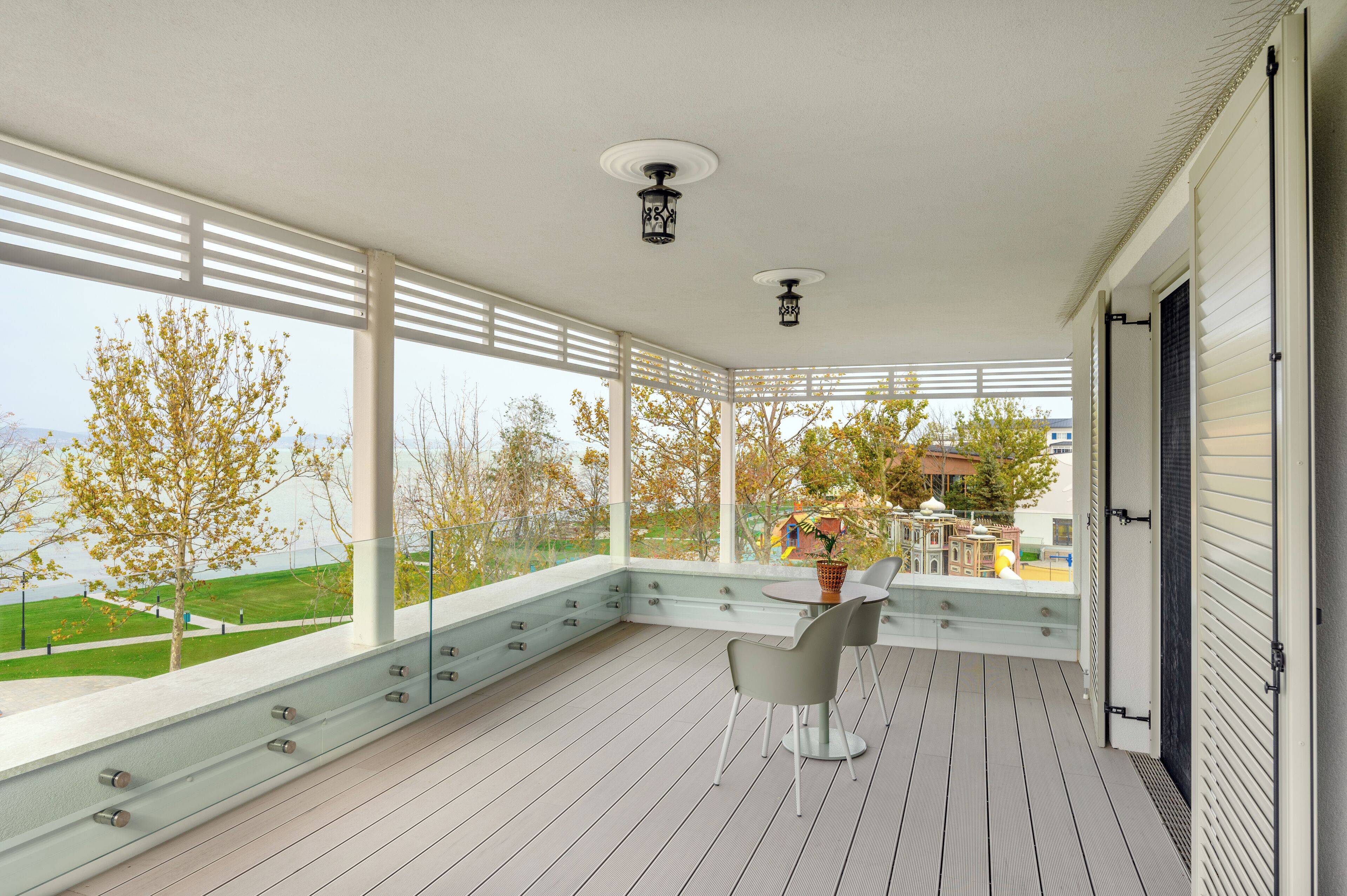 A covered balcony with a table and chairs, large windows, and a view of a park and buildings.