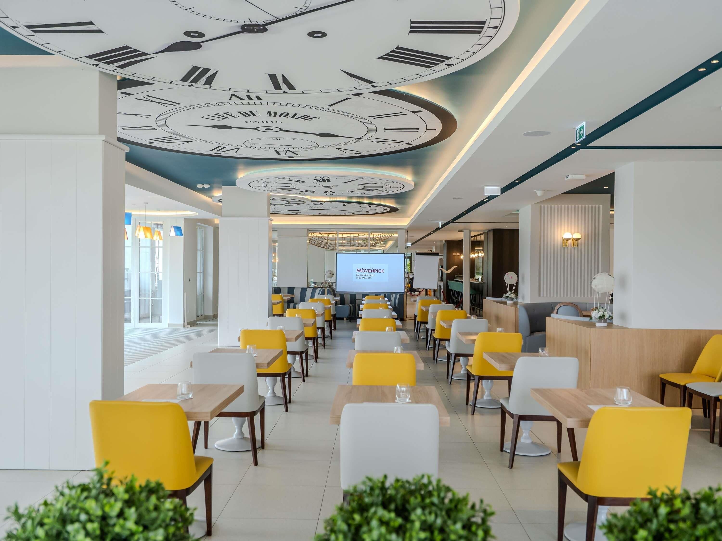 A modern restaurant with yellow and white chairs, a ceiling with clock designs, and a projection screen displaying 'MÖVENPICK'.