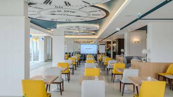 A modern restaurant with yellow and white chairs, a ceiling with clock designs, and a projection screen displaying 'MÖVENPICK'.