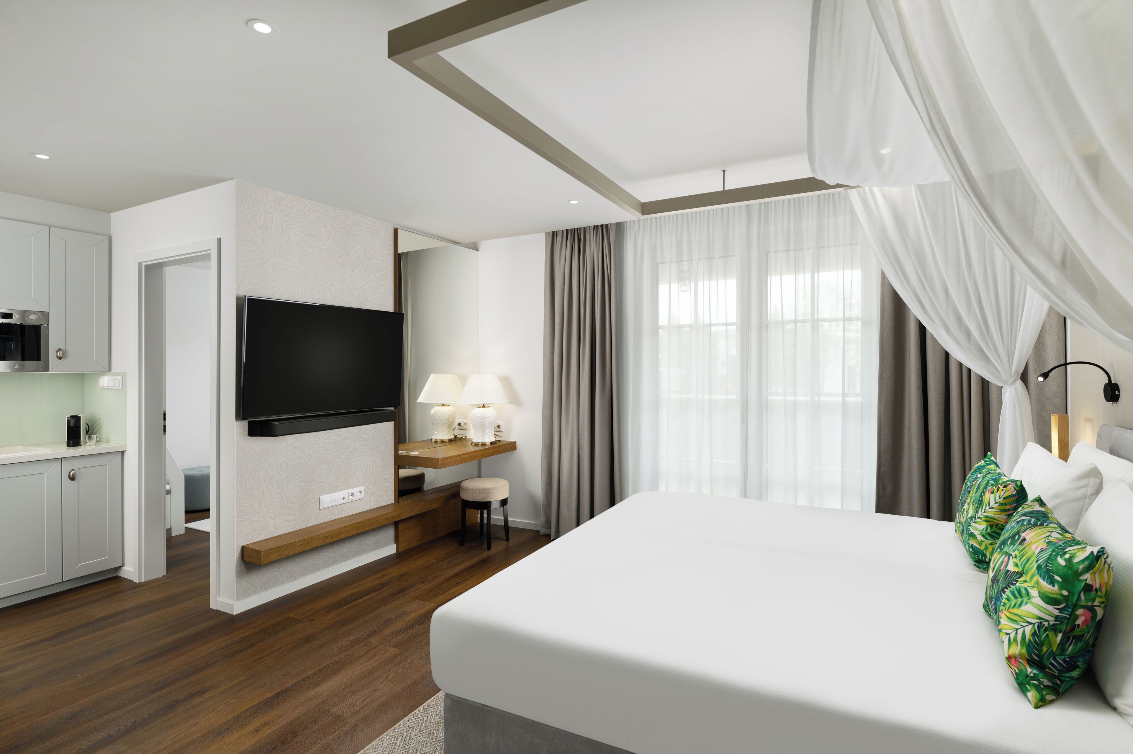 Quarto de hotel com cama, televisão, escrivaninha com luminária e cozinha compacta.