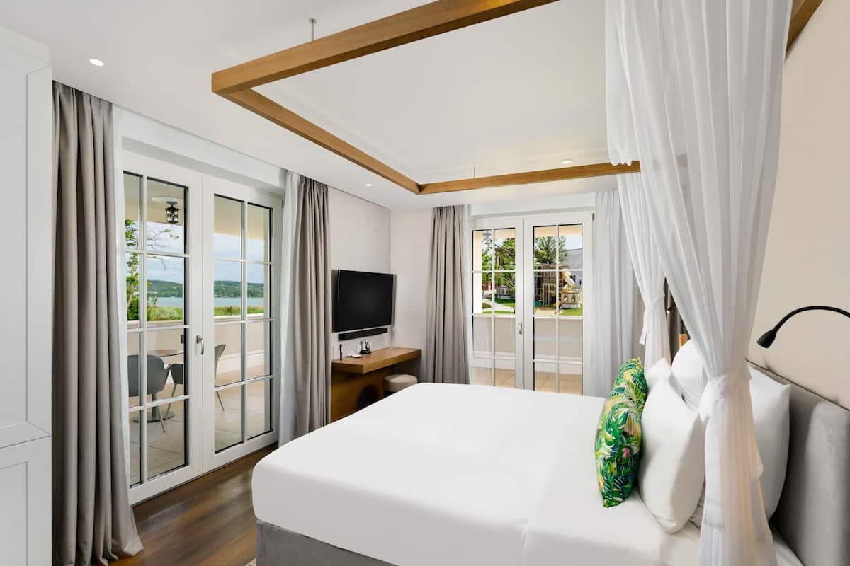 Habitación de hotel con una cama grande, televisión, balcón con cortinas y vista a la vegetación.