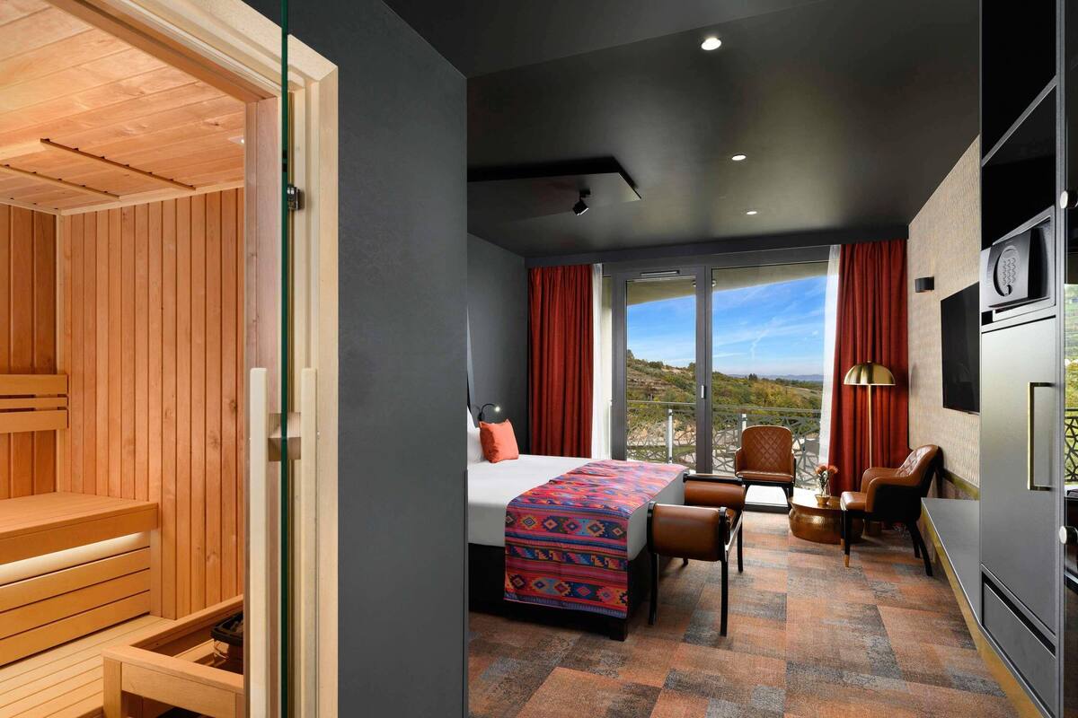 A modern hotel room with a wooden sauna, a bed with a colorful blanket, a desk with chairs, and a view of the outdoors.