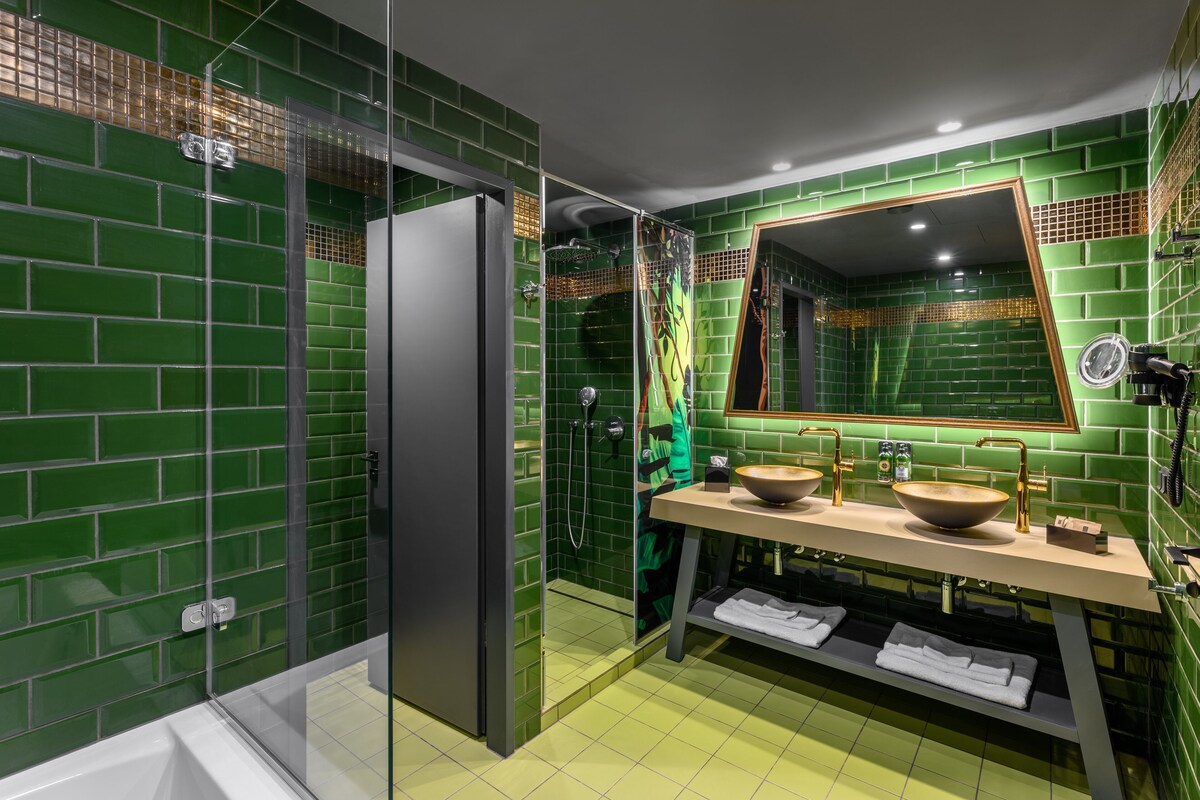 Une salle de bain moderne avec des murs recouverts de carreaux verts, une douche à parois de verre, un grand miroir et un meuble-lavabo beige doté d’un vasque.
