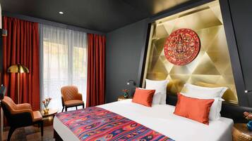 A modern hotel room with a large bed, red curtains, a decorative wall piece, and a small table with a lamp.