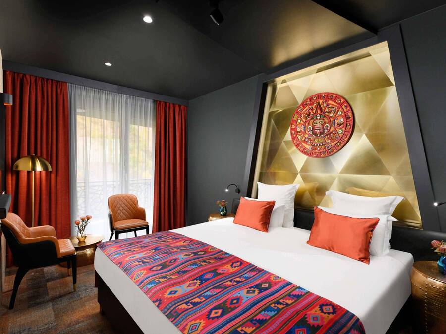 A modern hotel room with a large bed, red curtains, a decorative wall piece, and a small table with a lamp.