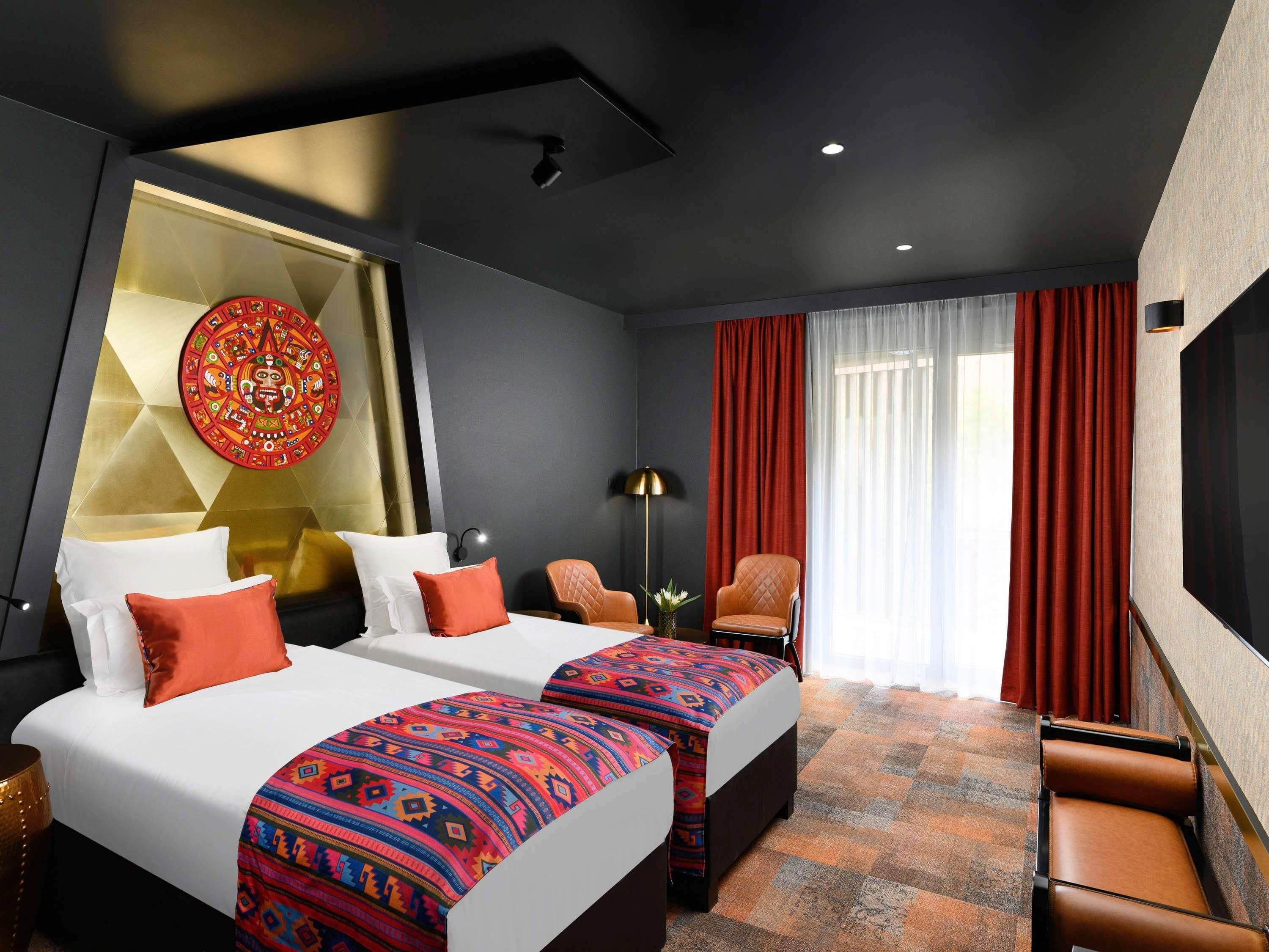 Quarto de hotel com duas camas, um grande painel decorativo, cortina vermelha e vista para o exterior.