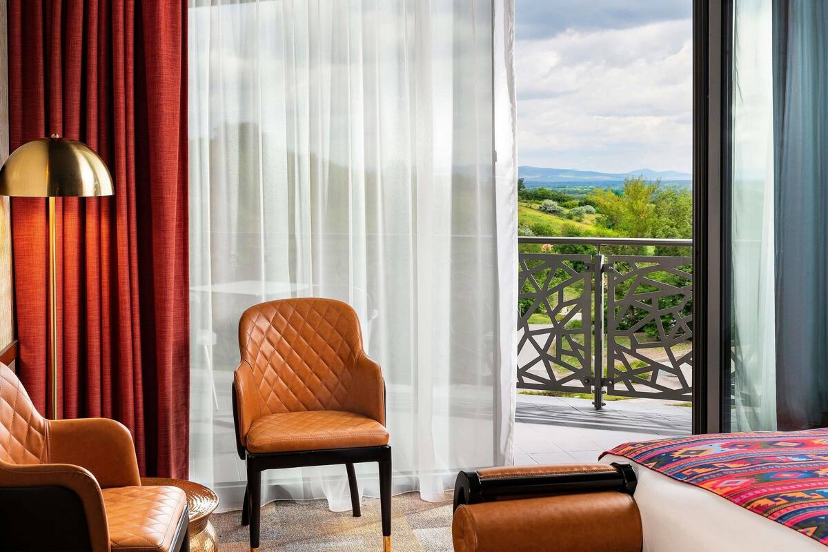 A hotel room with a bed, a brown leather chair, a small table, and a view of greenery through the window.