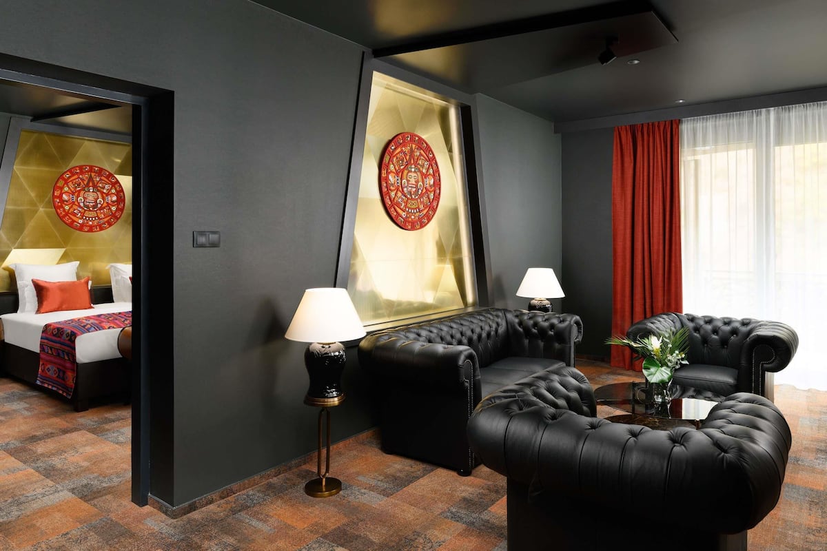 A modern hotel room with a bed, two black leather sofas, a large mirror, and a red curtain.
