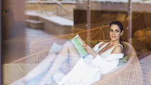 A person lying on a wicker lounge chair reading a book in a well-lit outdoor area with a stone wall and seating in the background.