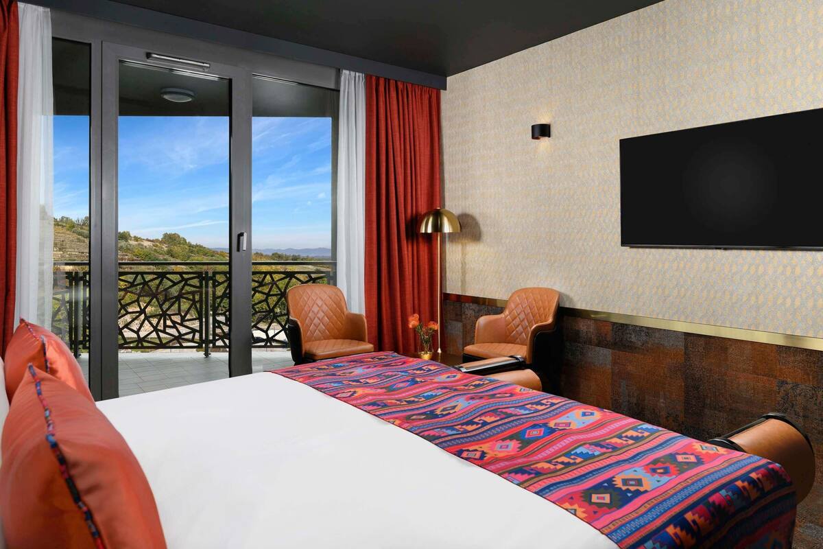 A hotel room with a bed, two chairs, a TV, and a balcony with a view.