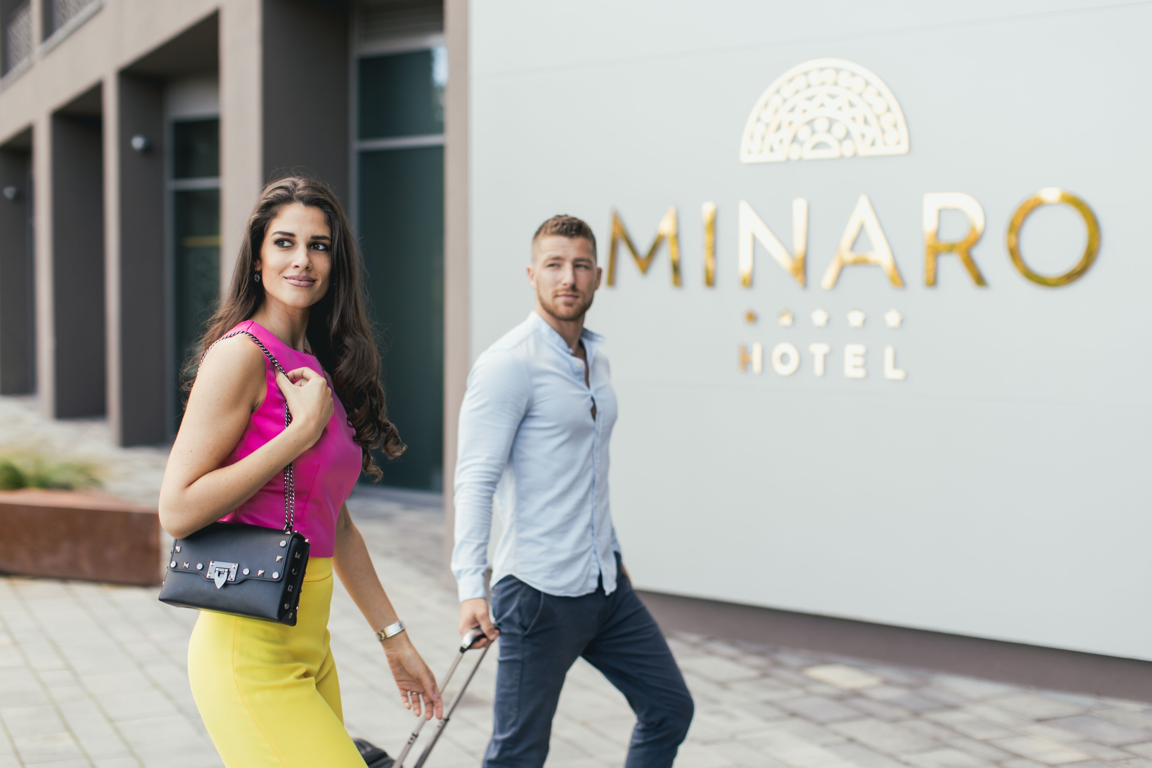 Two people walking past a hotel sign with the name "MINARO HOTEL" and a logo.