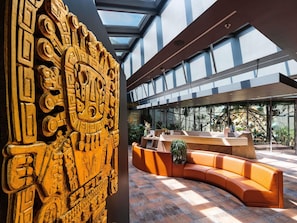 A modern lobby with a large wooden carving, a curved orange sofa, and a glass ceiling. - Minaro Hotel Tokaj - MGallery Collection (Tokaj)