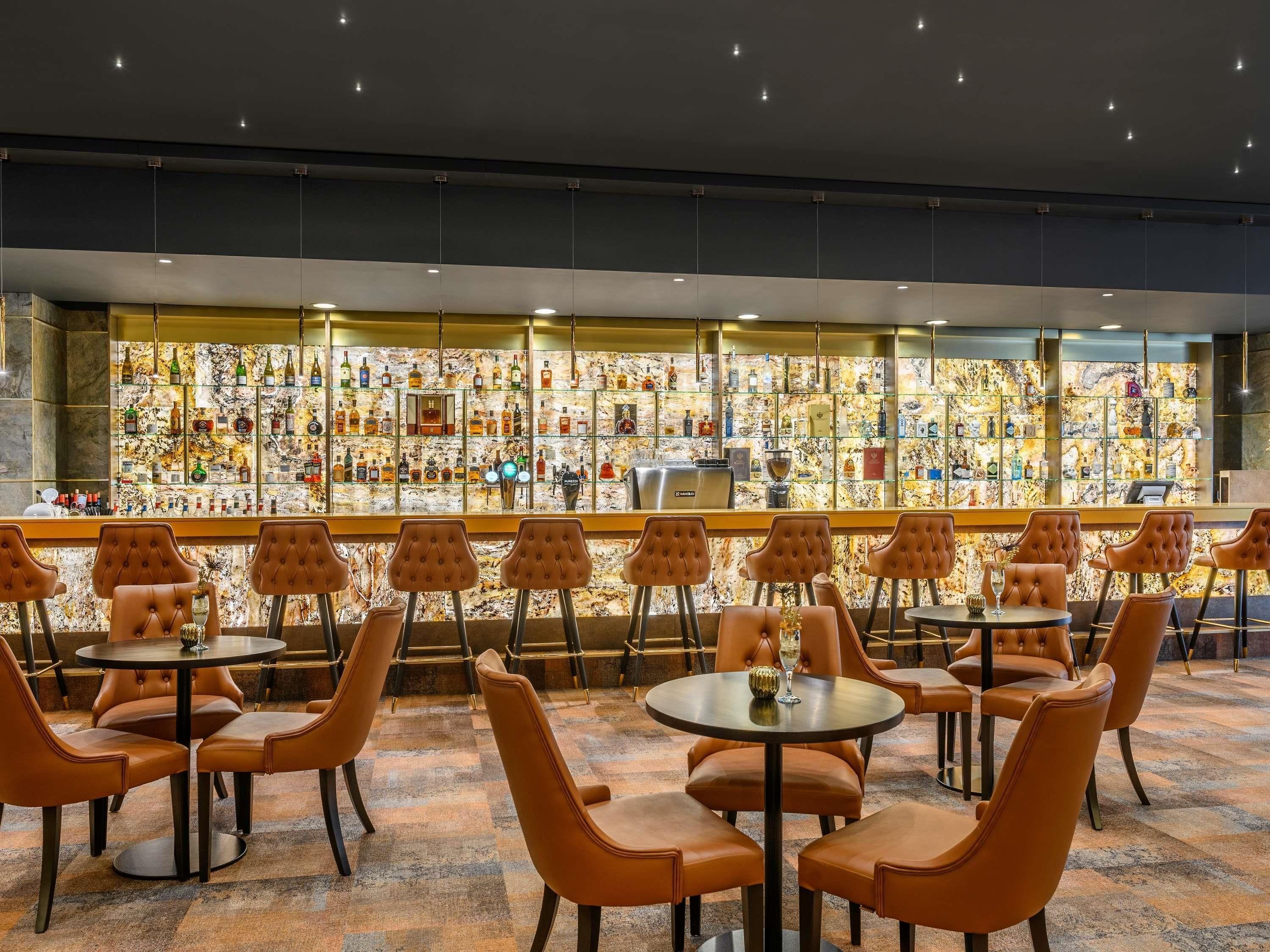 A bar area with a marble wall and a variety of liquor bottles. There are several round tables and chairs arranged for seating.