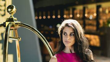 Een vrouw in een roze top en gele broek leunt tegen een gouden balustrade.